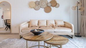 Een woonkamer met een beige bank, twee houten salontafels, een decoratieve wandversiering en een vloerlamp.