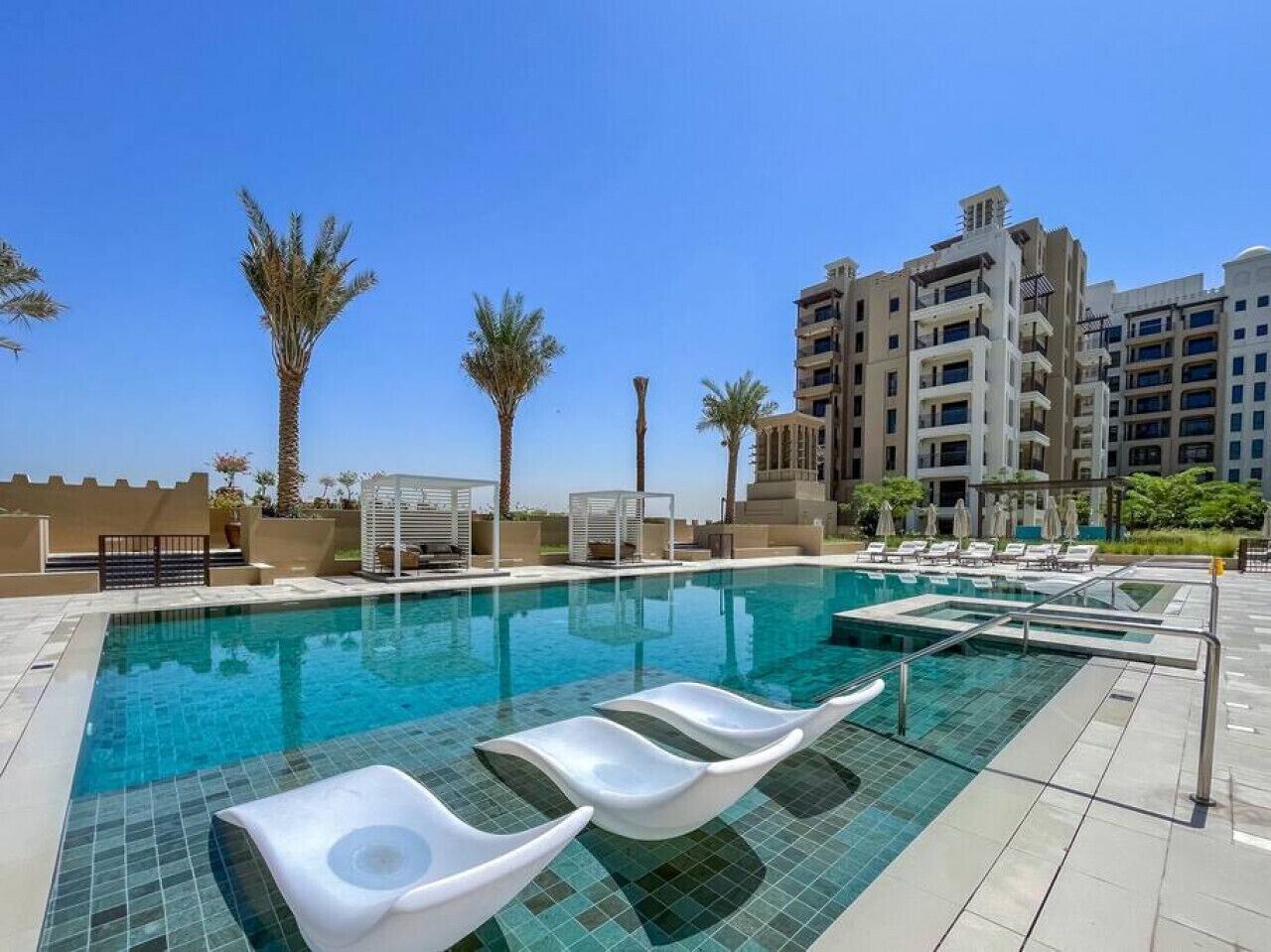 A swimming pool with lounge chairs, palm trees, and modern buildings in the background.