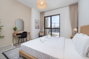 A modern hotel room with a large bed, a desk, a chair, a large window with a view of buildings, and a hanging light fixture.