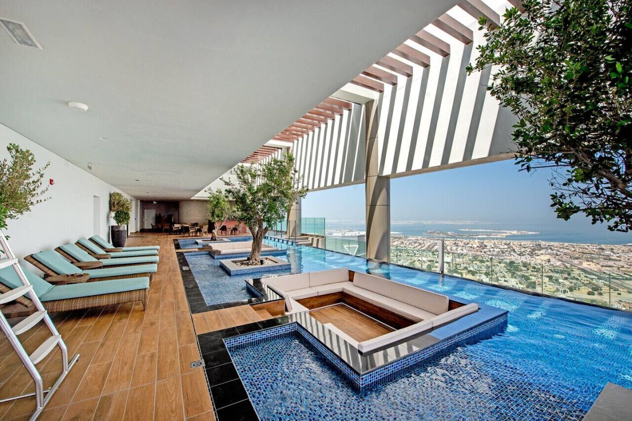 A rooftop infinity pool with lounge chairs, a wooden deck, and a view of the city.