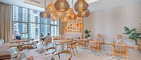 Un espace de restauration moderne doté de nombreuses tables et chaises, de grandes fenêtres offrant une vue sur la ville et de luminaires décoratifs en fibres tressées.
