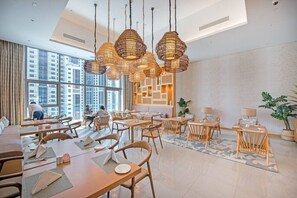 Un comedor moderno con varias mesas y sillas, ventanas amplias con vista a la ciudad, y lámparas decorativas de fibra.