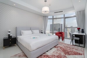 Habitación de hotel moderna con una cama grande, un escritorio con una silla, una silla roja y vistas de la ciudad a través de amplios ventanales.
