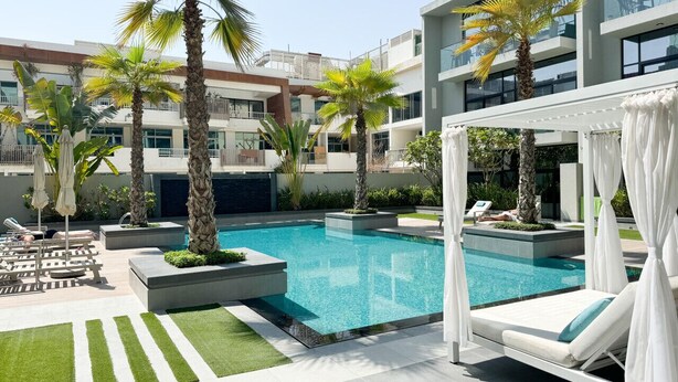 Un área de piscina moderna con sillones de descanso, palmeras y una zona de estar cubierta.