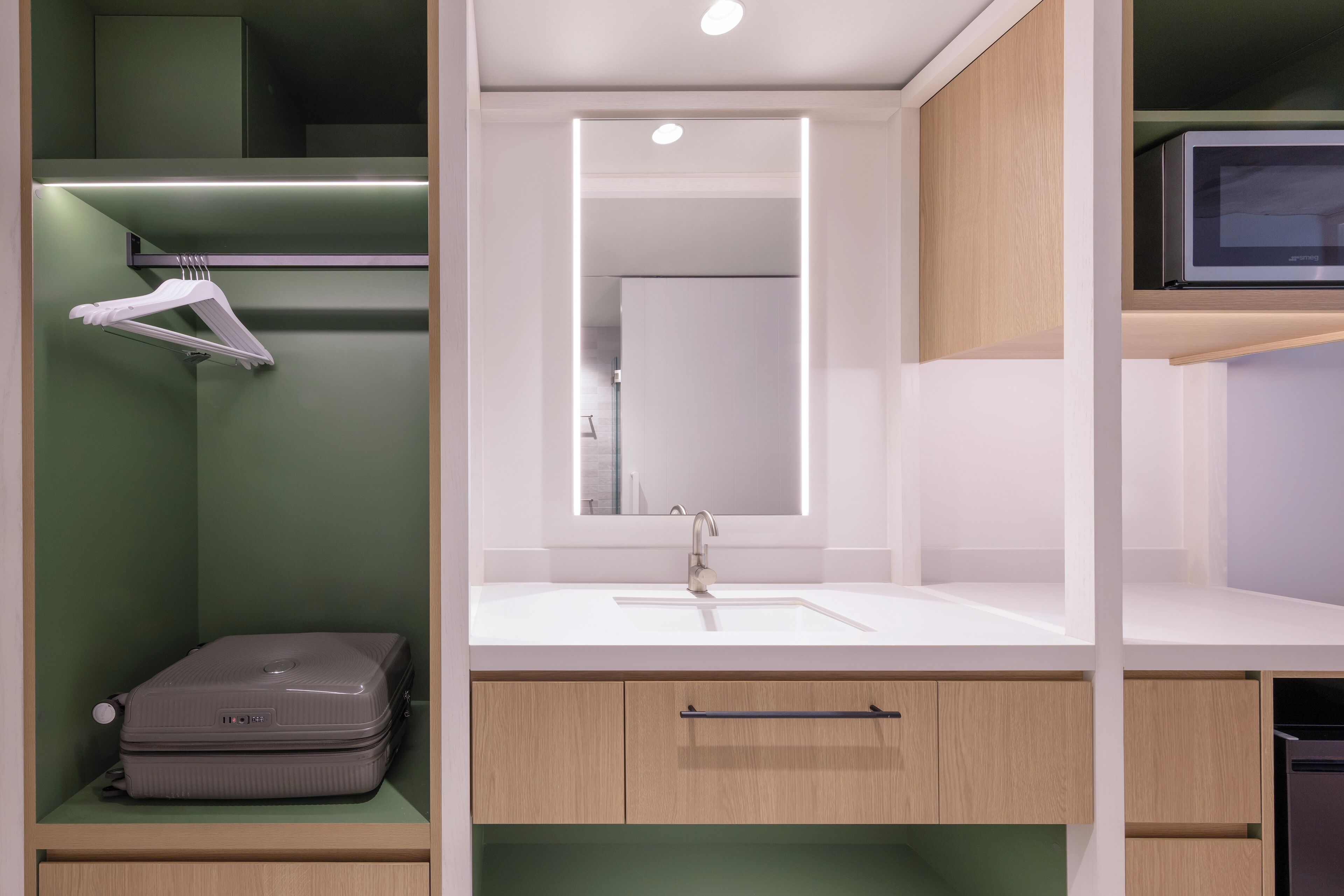 Un baño moderno con un lavabo blanco, un espejo y una maleta sobre una estantería.
