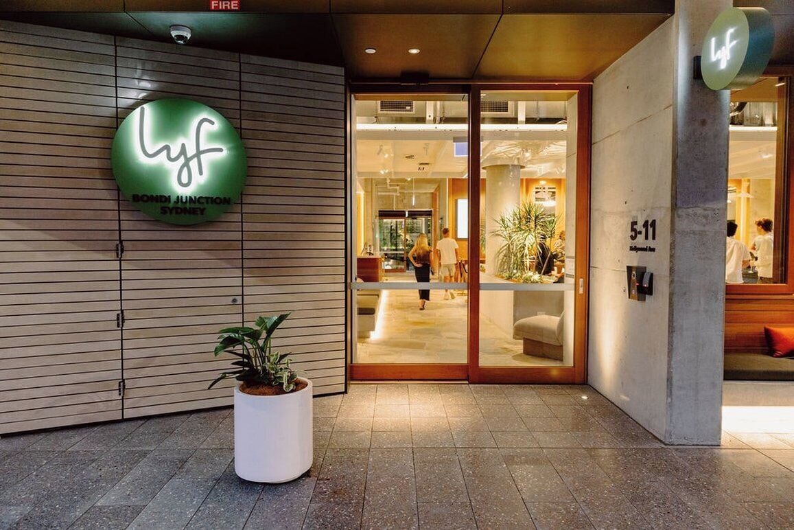 Entrada de un hotel con un letrero que dice 'Lyf Bondi Junction Sydney', una planta en maceta y un detector de incendios.