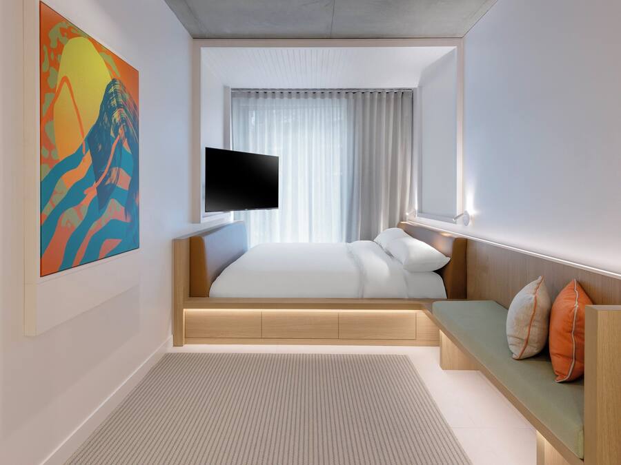A modern hotel room with a bed, a flat-screen TV, a bench with pillows, and a large abstract painting on the wall.
