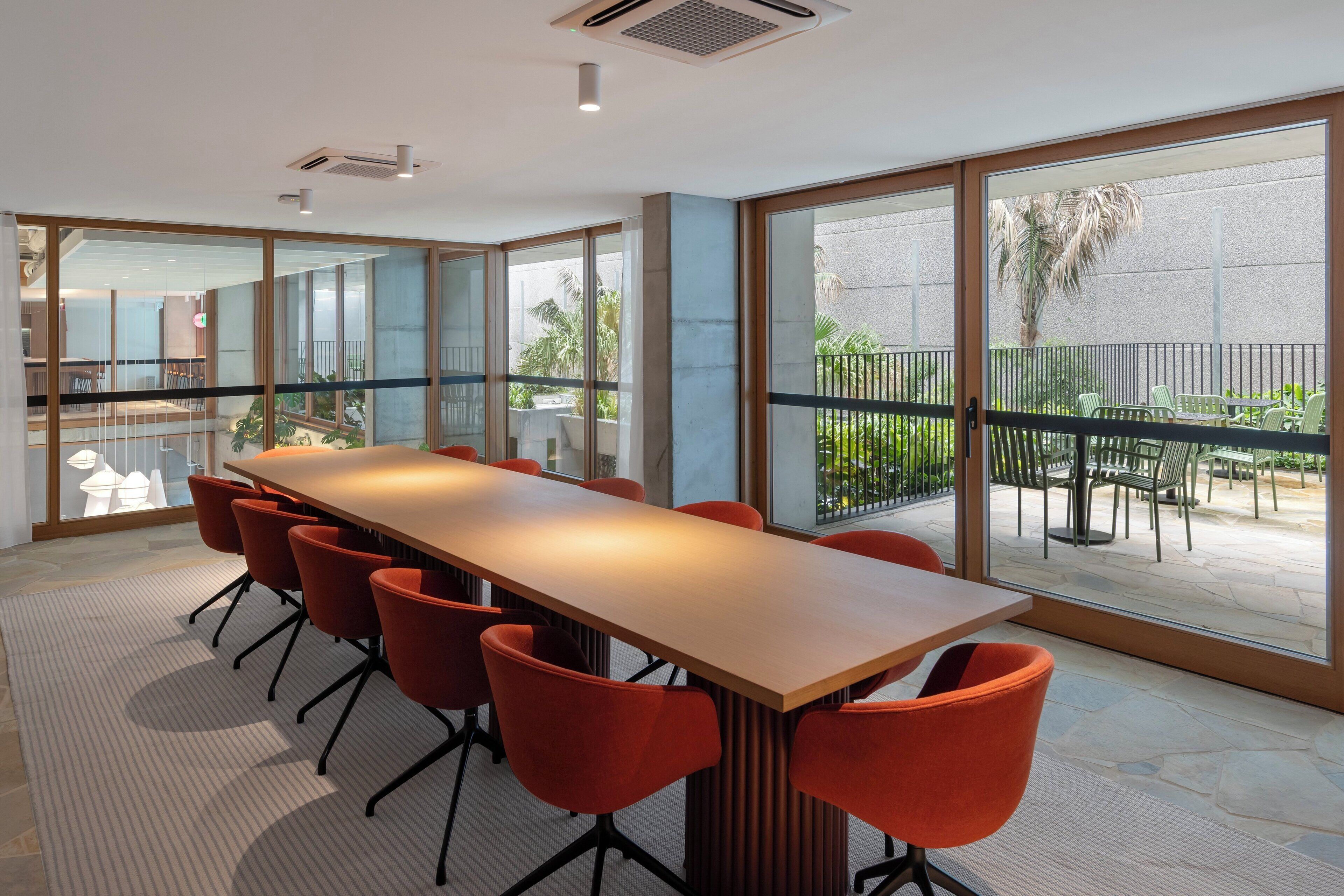 A conference room with a long table and orange chairs, large windows, and a view of a balcony with outdoor furniture.