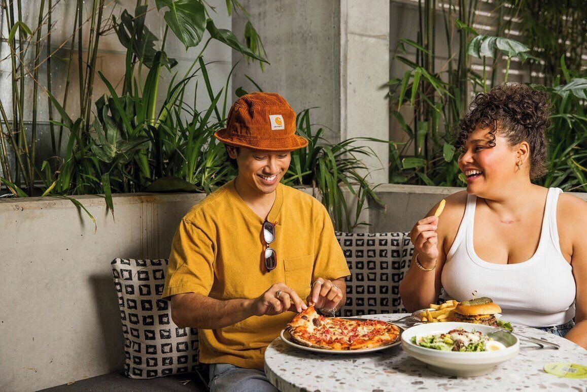 Deux personnes dînent à une table en plein air, devant une pizza et une salade, entourées de plantes et d’un coussin à motifs.