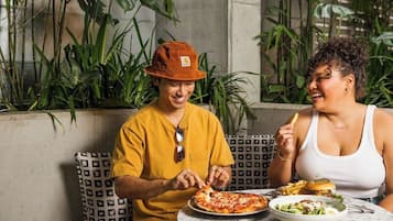 Deux personnes dînent à une table en plein air, devant une pizza et une salade, entourées de plantes et d’un coussin à motifs.