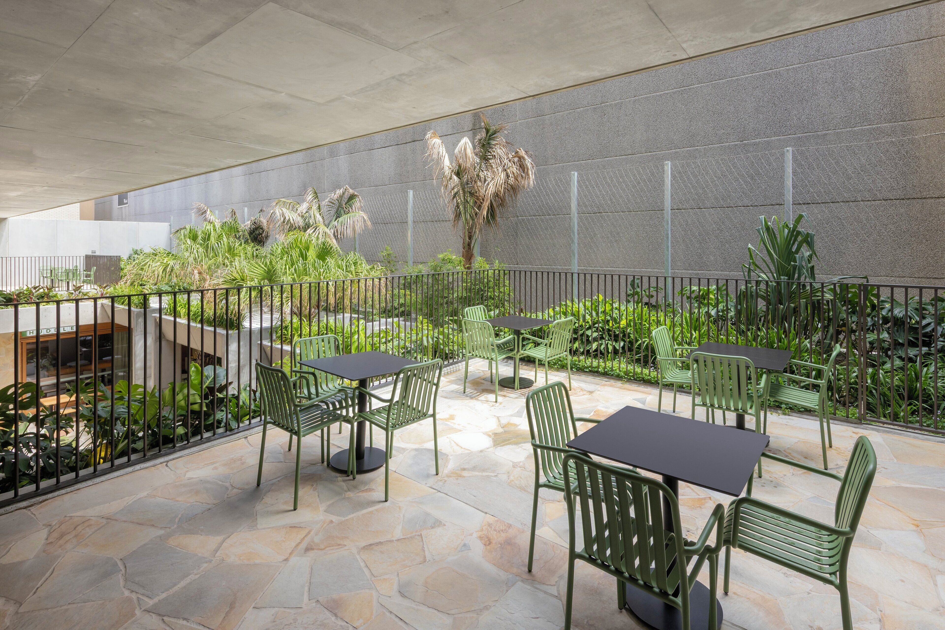 An outdoor patio area with stone flooring, green metal chairs and tables, and a variety of plants.