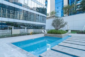Un área de piscina exterior moderna con una piscina rectangular, rodeada por un muro blanco bajo y un edificio de fachada de vidrio al fondo.