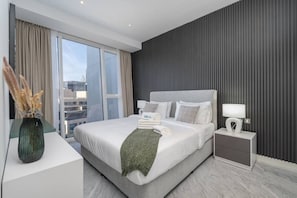 A modern hotel room with a large bed, a nightstand, a lamp, and a view of the city through the window. - The Pad 1109 - Business Bay · Cosmopolitan Apartment with Burj Khalifa View (Dubai)
