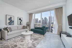 Ein modernes Wohnzimmer mit einer Couch, einem grünen Sessel und einem Couchtisch. An den Wänden befinden sich zwei abstrakte Gemälde, ein großes Fenster mit Vorhängen und Blick auf die Stadt.