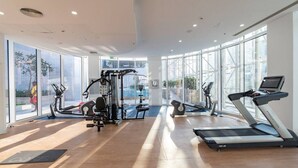 A modern gym with cardio machines, weightlifting equipment, and large windows offering views of the outdoors. - The Pad 1109 - Business Bay · Cosmopolitan Apartment with Burj Khalifa View (Dubai)