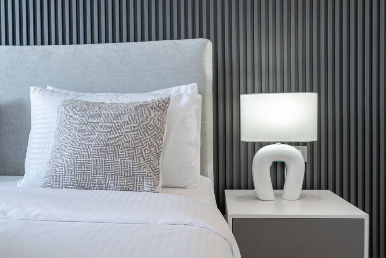 A neatly made bed with white bedding and patterned pillows, a modern bedside table with a white lamp, and a wall with vertical slats.