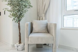 Una sala de estar moderna con un sillón beige, una planta grande en maceta y vistas de un paisaje urbano a través de un ventanal.
