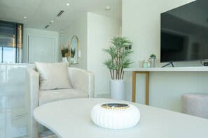 Una sala de estar moderna con un sofá blanco, una mesa blanca con un cuenco decorativo, un televisor de pantalla plana grande y una planta en maceta.
