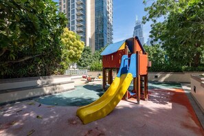 Un parque infantil con un tobogán y estructuras para escalar, rodeado de árboles y edificios.