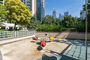 Un área de ejercicios al aire libre con bicicletas estáticas y una pequeña piscina, rodeada de follaje y edificios altos al fondo.