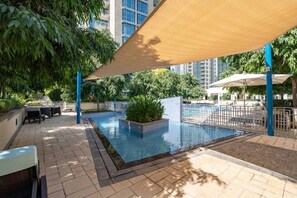Una piscina al aire libre con solárium de baldosas, rodeada de follaje y una zona sombreada.