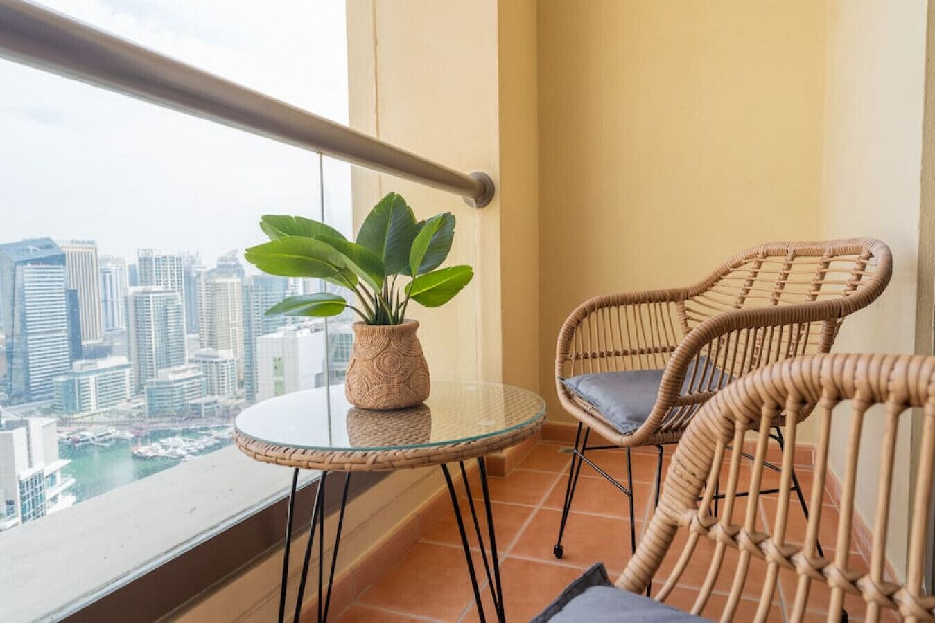 Un balcon avec une table en verre, une chaise en osier, un pot de fleurs et une vue sur le paysage urbain.