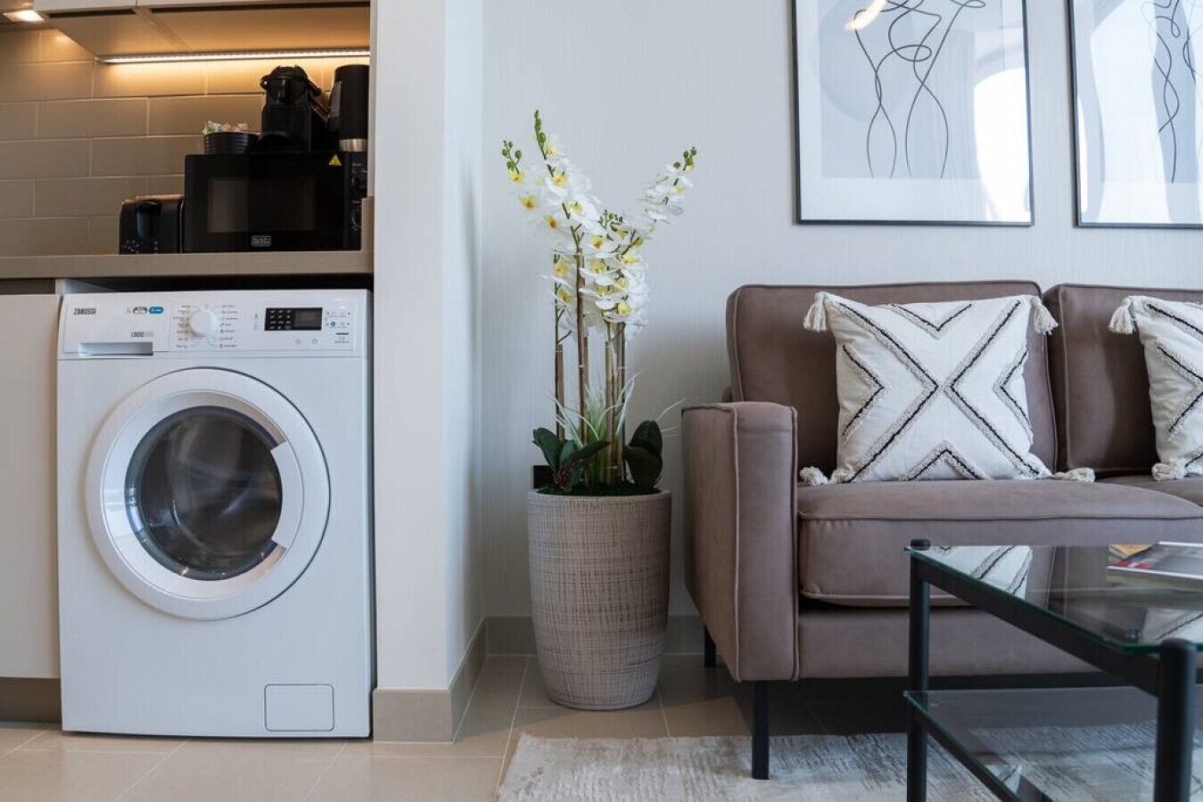 Un espace de vie compact comprenant un lave-linge à chargement frontal, un canapé avec des coussins à motifs, une plante en pot et des œuvres d’art abstraites accrochées au mur.