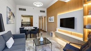 A modern living room with a TV, sofa, dining table, and kitchen in the background. - Marina Breeze 1BR Walk to Beach (Dubai)