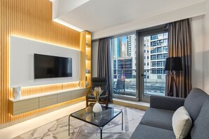 Uma sala de estar moderna com uma TV de tela plana, um sofá, uma mesa de centro de vidro e vista para a paisagem urbana através de grandes janelas.