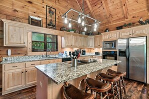 A kitchen with wooden cabinets, granite countertops, a stainless steel refrigerator, and a bar stool area. - Mini Golf |Games| Fire Pit| Mountain Medicine (Sevierville)