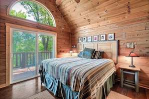 A cozy cabin bedroom with a large bed, a small table with a lamp, and a view through a sliding door to an outdoor deck.