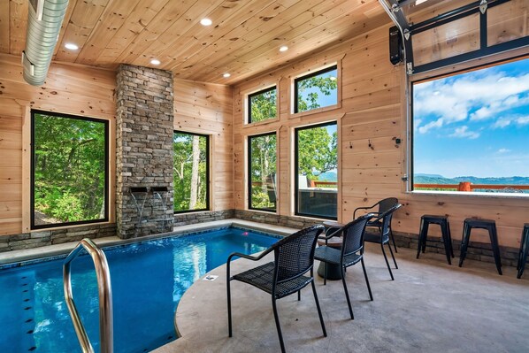 An indoor swimming pool with a stone fireplace, wooden walls, and large windows offering a view of the outdoors.