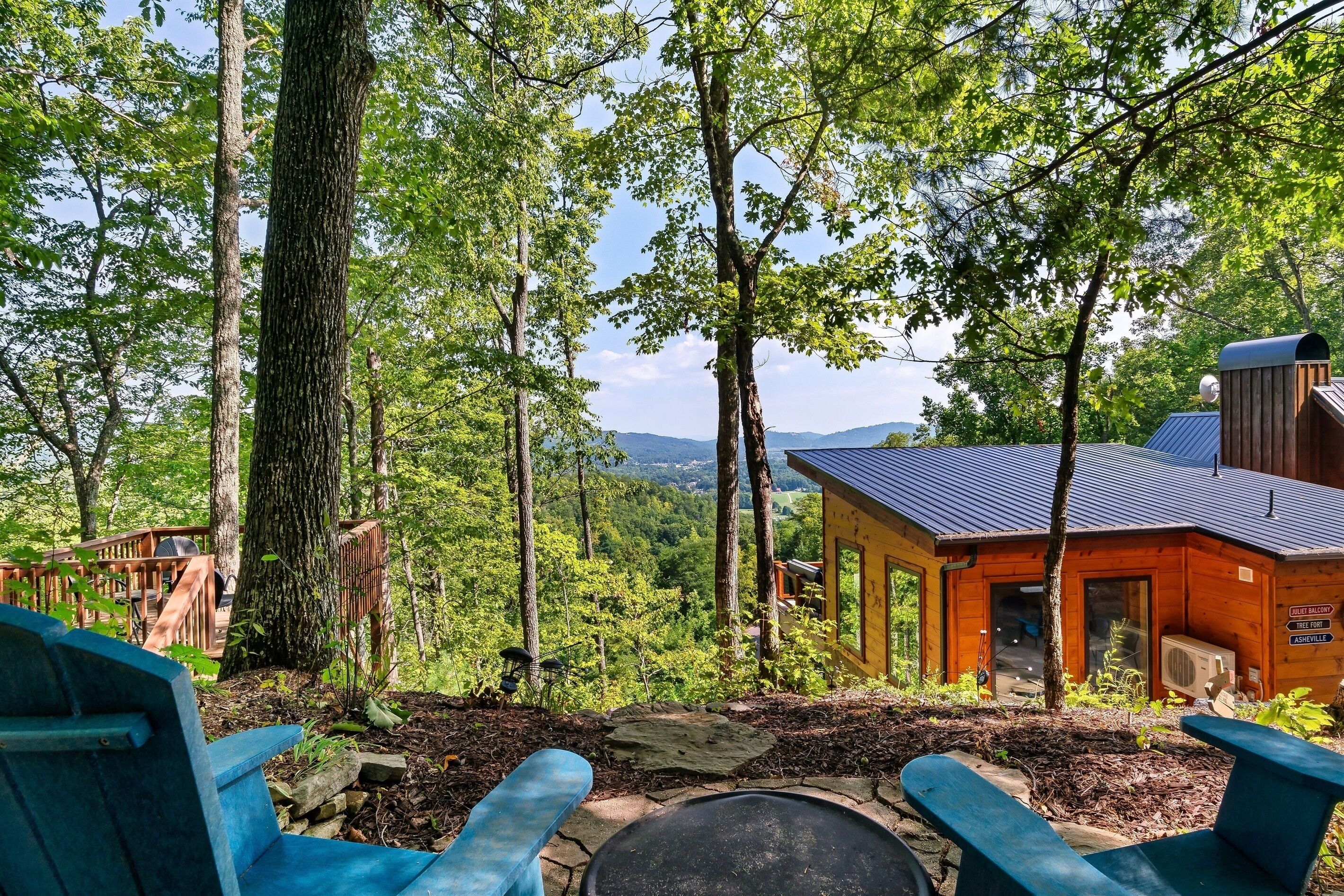 Een chalet met een vuurkuil van steen en blauwe Adirondack-stoelen, omgeven door bomen en uitzicht op een berg.