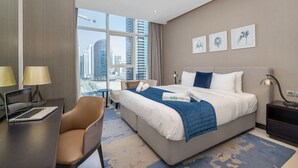 Habitación de hotel con una cama grande, un escritorio, una silla y vistas de la ciudad a través de amplios ventanales.