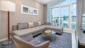 Una sala de estar moderna con un sofá seccional gris, una mesa redonda y una butaca. Hay cuadros enmarcados en la pared, una lámpara de pie y vistas de la ciudad a través de amplias ventanas.