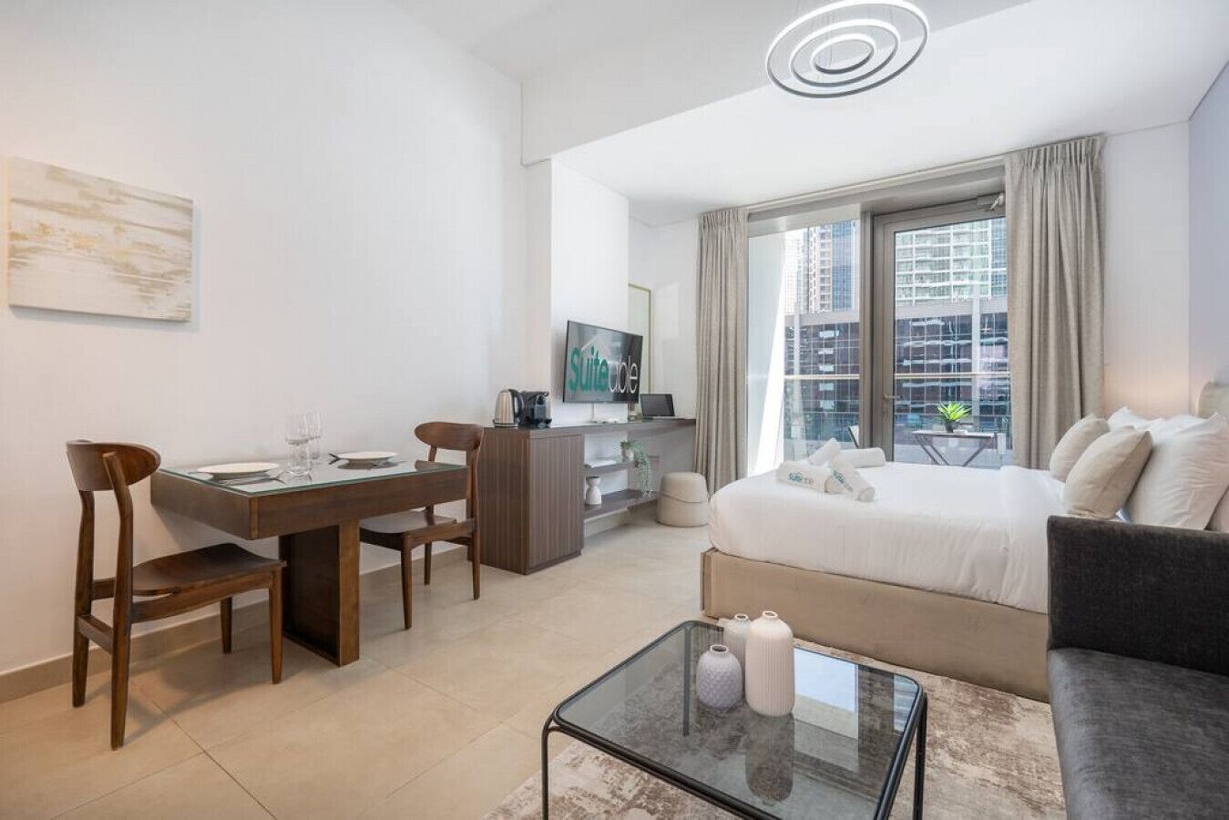 Een moderne hotelkamer met een bed, een sofa, een glazen salontafel, een eettafel met stoelen, een televisie en uitzicht op de stad door het raam.