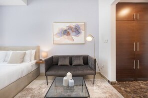 Une chambre d’hôtel moderne avec un lit, un canapé gris, une table basse en verre ornée de vases, un tableau au mur et une armoire.