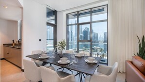 A dining area with a table set for four, large windows with a city view, a kitchen area, and a brown leather sofa. - 15 Northside - 210A · Contemporary Ranch Living with Burj Khalifa View (Dubai)