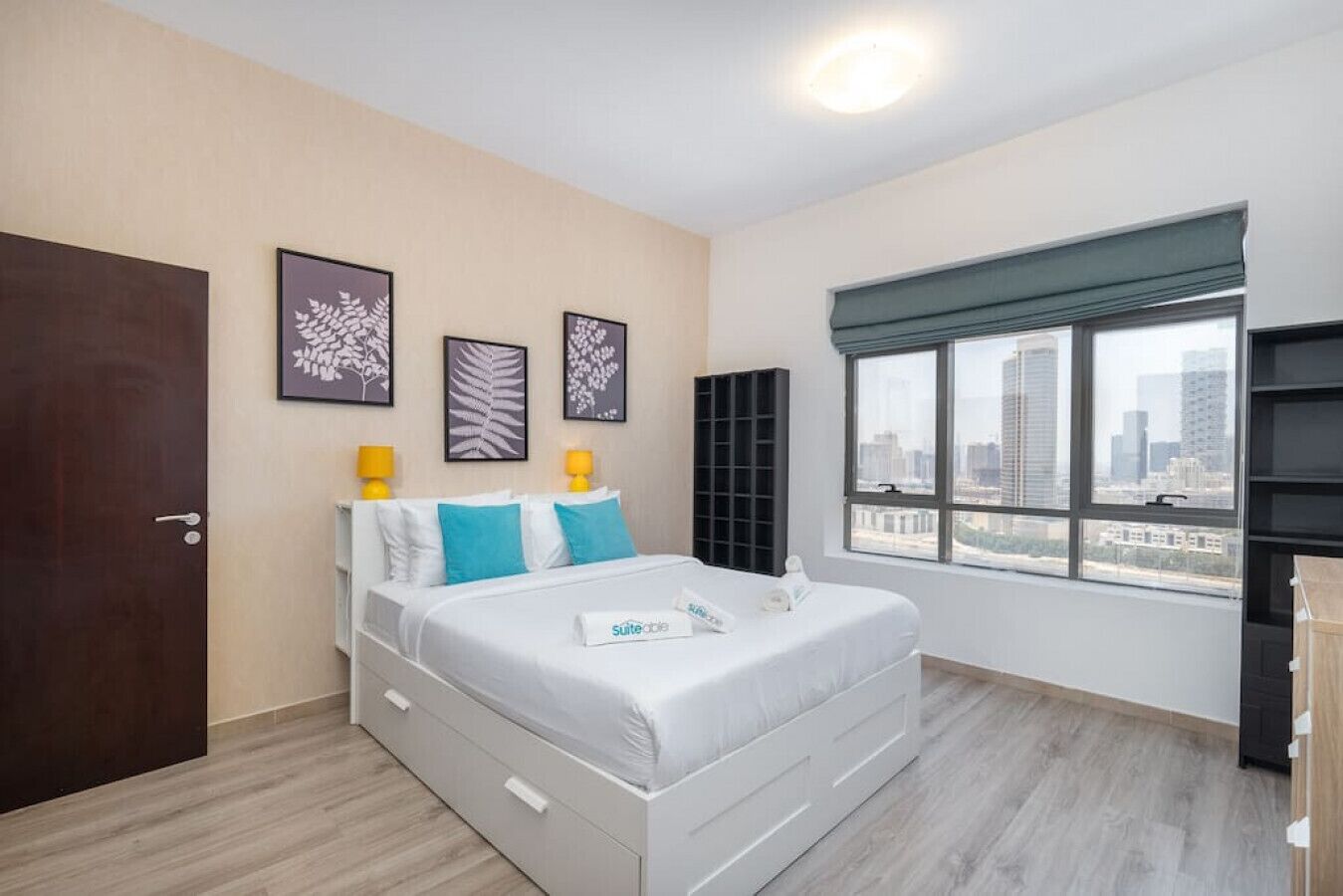 A hotel room with a bed, a window with a city view, a door, and wall-mounted artwork.