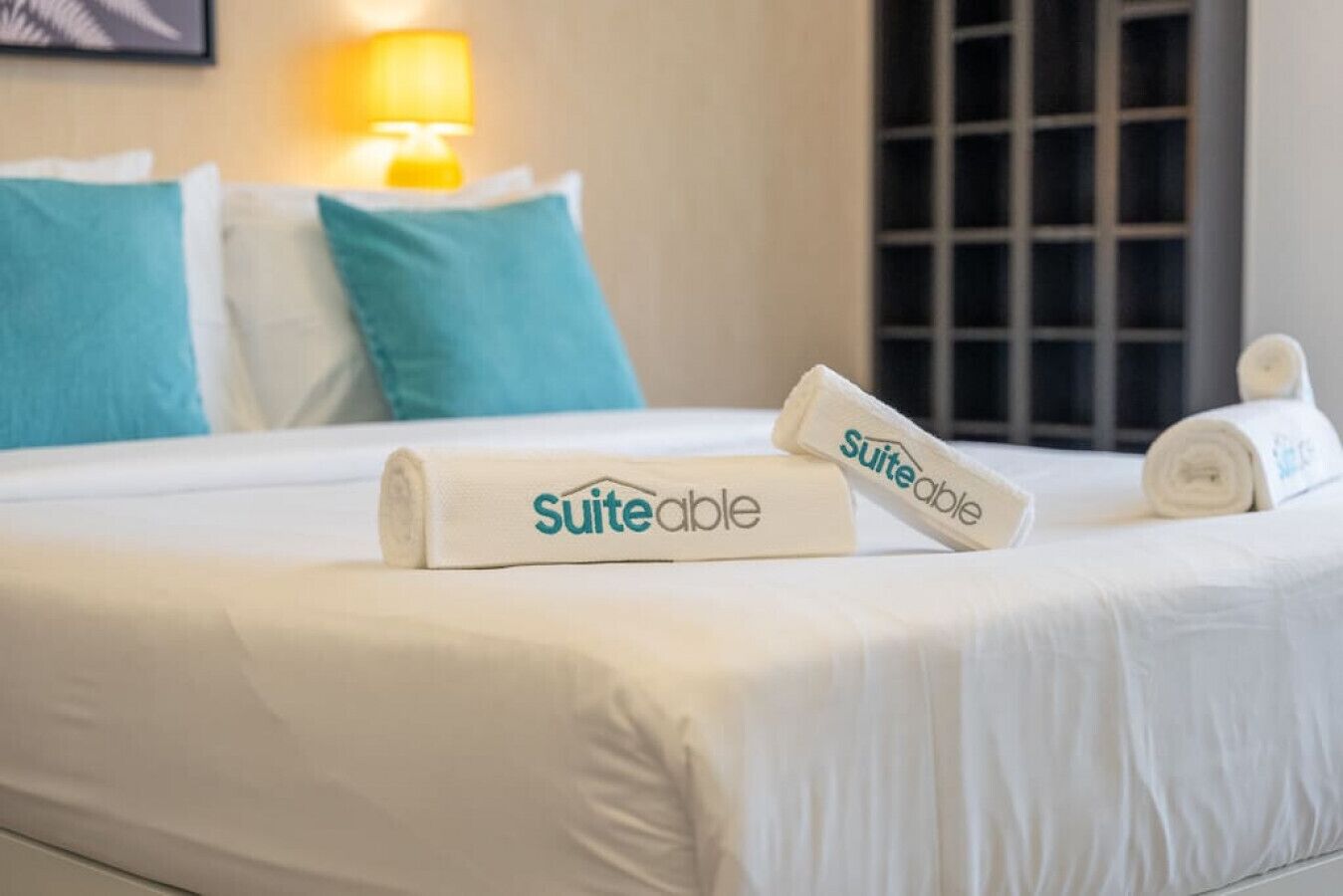 Ein ordentlich bezogenes Bett mit weißen Leintüchern und zwei zusammengerollten Handtüchern mit dem 'Suiteable'-Logo.