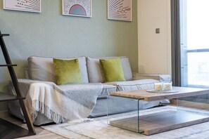 Een moderne woonkamer met een bank, groene kussens, een bijzettafeltje en een groot raam.