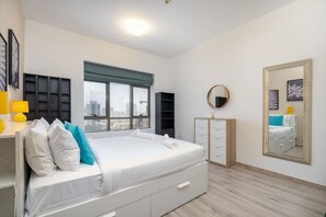 A modern bedroom with a large bed, a dresser, a wardrobe, a round mirror, and a city view through a window.