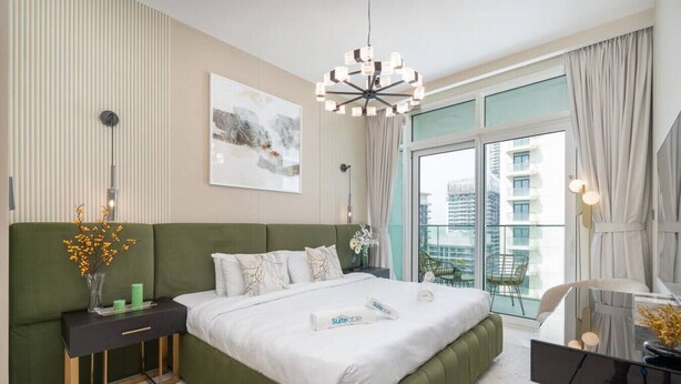 Une chambre moderne avec un lit recouvert d’un tissu vert, une table de chevet ornée d’un vase de fleurs, une grande fenêtre avec des rideaux et une vue sur la ville.