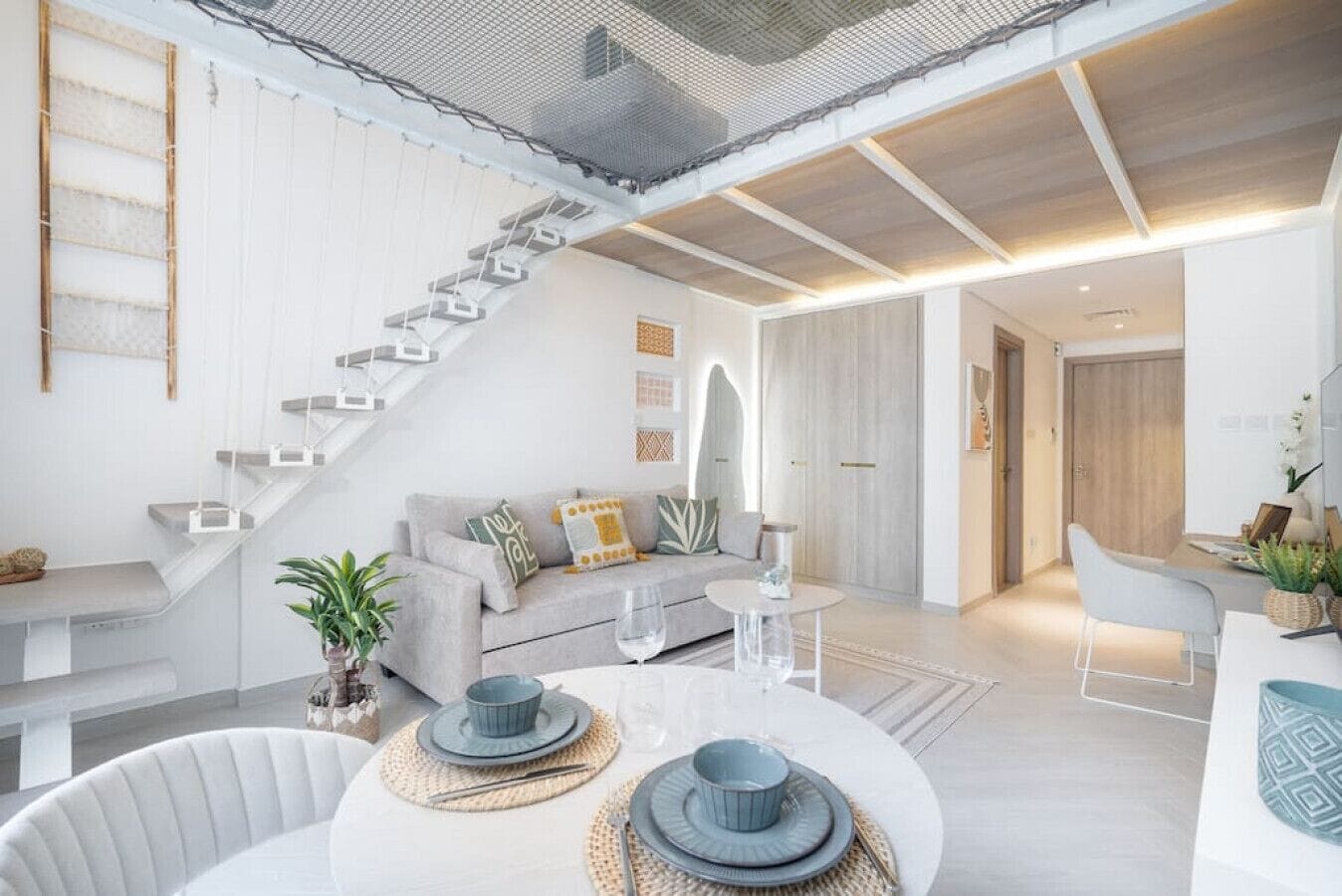 A modern living room with a staircase, a dining area with a round table, and a kitchen area.