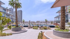 Uma área de piscina no terraço com espreguiçadeiras, palmeiras e edifícios modernos ao fundo.