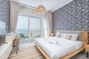 A modern hotel room with a large bed, a desk, and a balcony with a view of the sea.