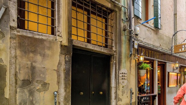 Edifici històric amb una porta negra, una petita zona d'seients a l'exterior i un rètol de botiga visible.