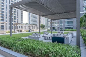 A covered outdoor seating area with modern furniture, surrounded by greenery and high-rise buildings in the background.