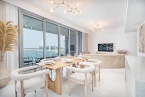 A modern dining area with a wooden table set for six, white cushioned chairs, and a large window offering a view of the sea. - Your beachfront luxe escape experience - Suiteable (Dubai)