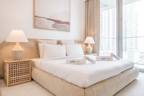 A hotel room with a large bed, two bedside tables with lamps, a view of tall buildings through large windows, and a piece of abstract art on the wall.